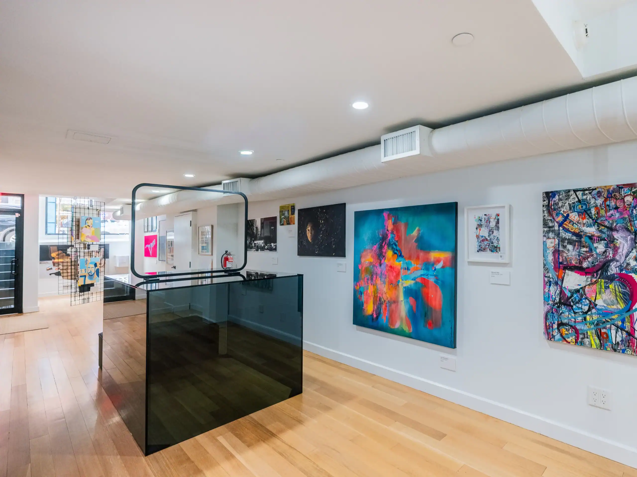 A modern art gallery with light wood floors and white walls displays various colorful abstract paintings. A glass railing surrounds a stairwell in the center of the room. Ceiling lights illuminate the artwork.