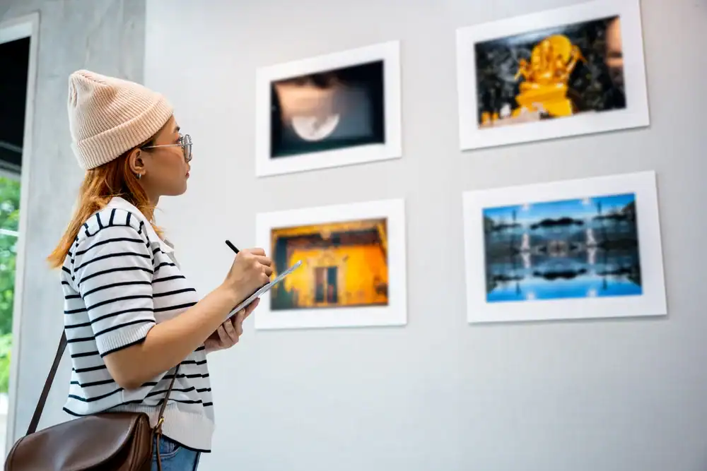 A person wearing a beanie, glasses, and striped shirt holds a notebook and pen, observing framed photographs displayed on a gallery wall.