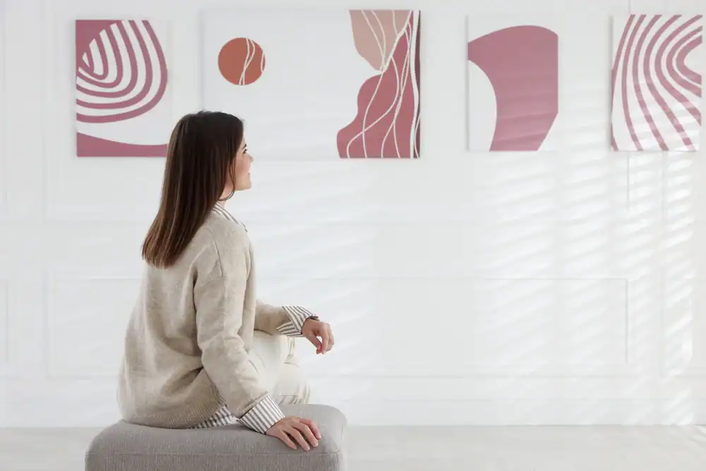 A woman with long brown hair sits on a gray ottoman, looking at three abstract pink and white paintings on a white wall in a bright, modern room.