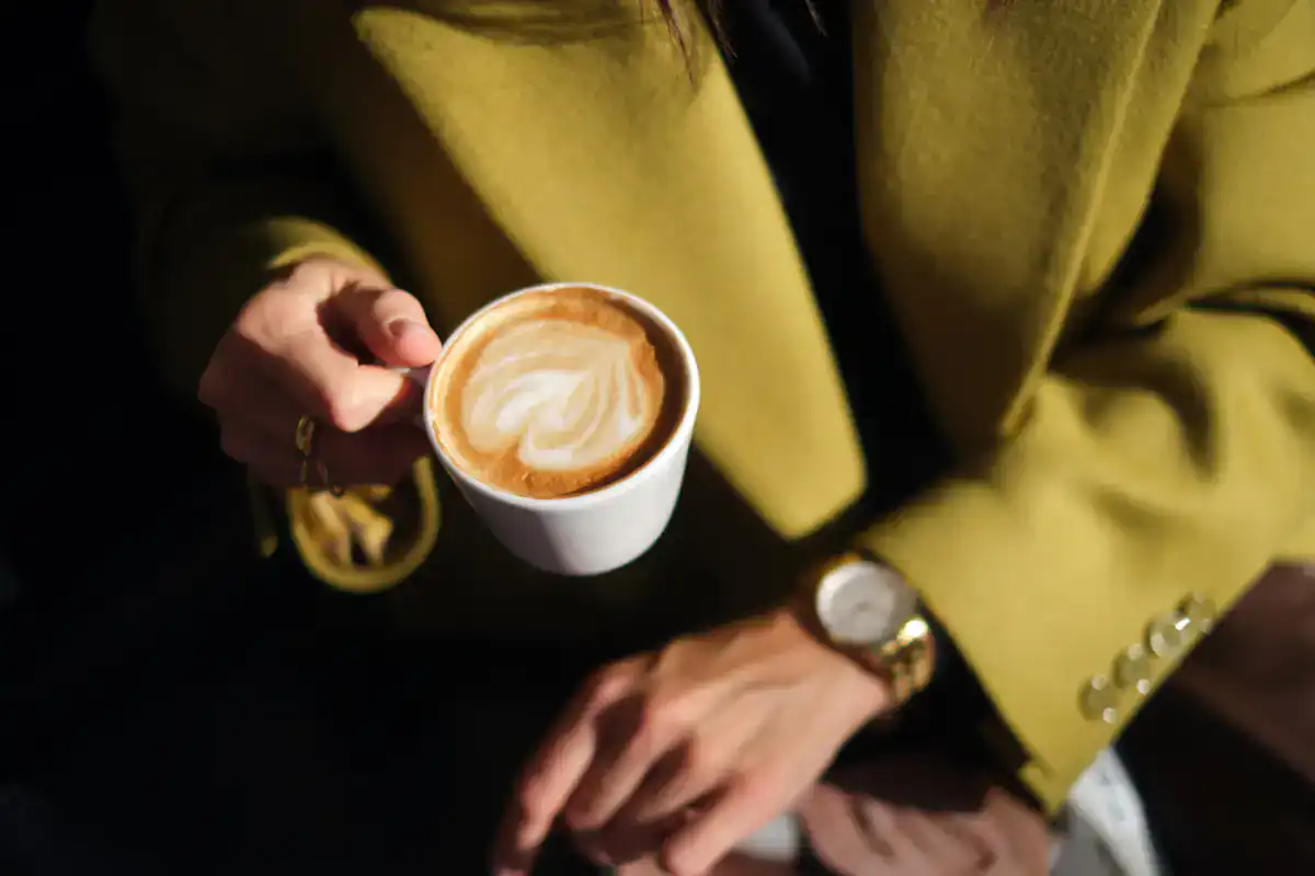 A person wearing a yellow-green coat holds a cappuccino with latte art at an Art Gallery Cafe in NYC