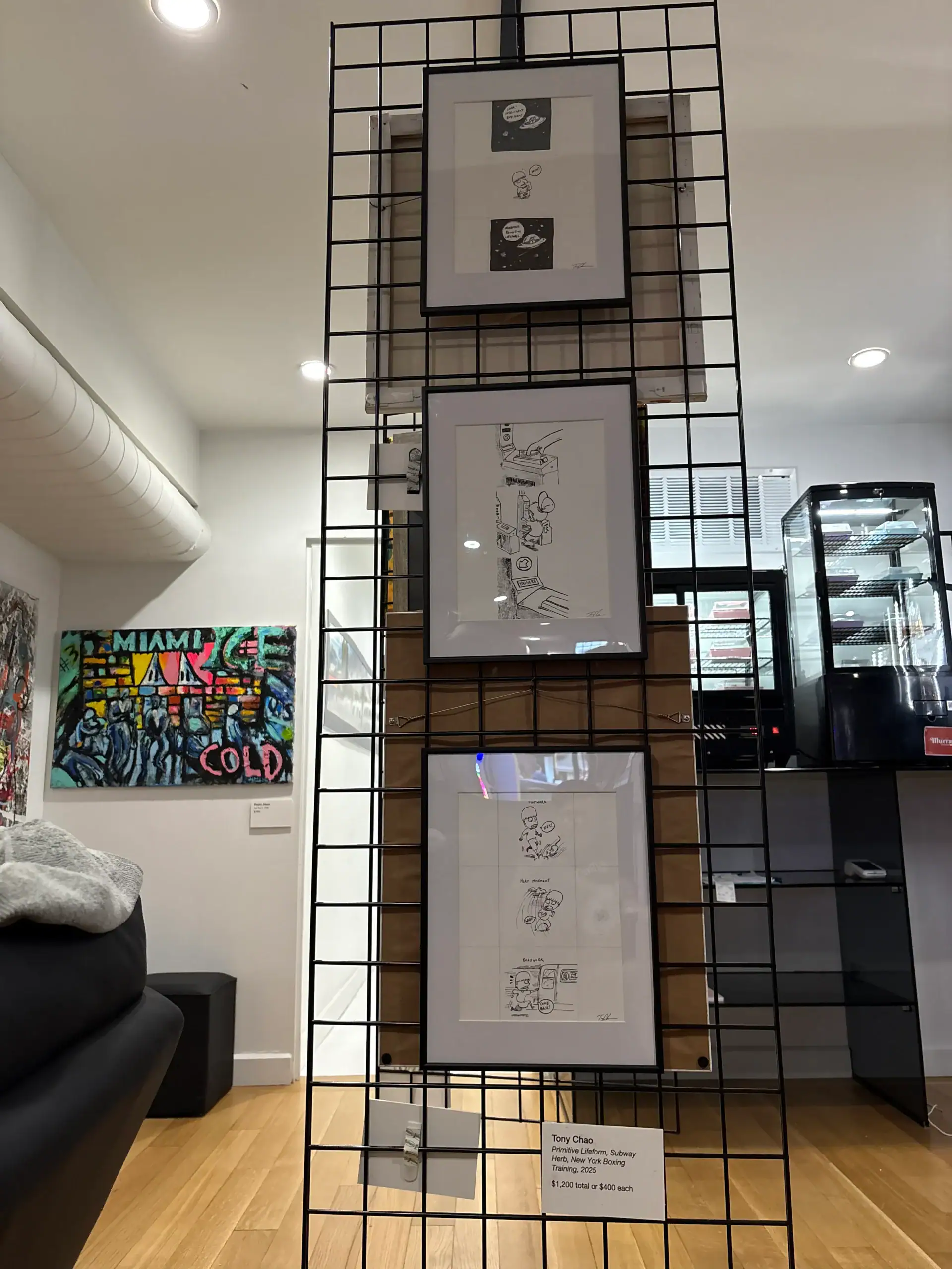 Three black-and-white framed drawings hang vertically on a metal grid in a NYC wine bar and art gallery.