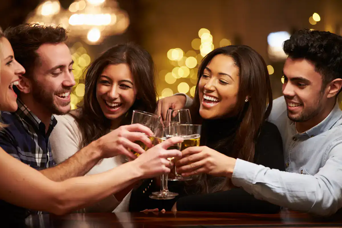 Five friends smile and clink glasses at a NYC Wine Bar & Art Gallery, with warm lights glowing behind.